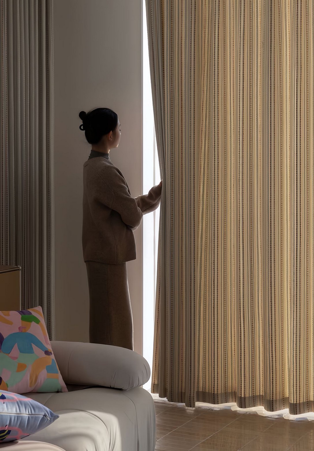 A person standing by a window, pulling back a semi-sheer, striped curtain in a living room.