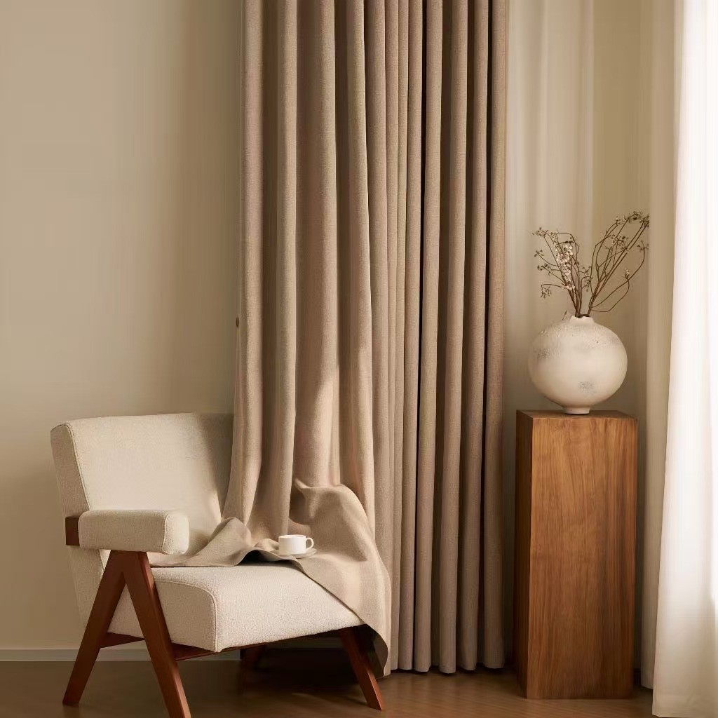 A beige armchair next to a wooden pedestal with a white vase, with floor-to-ceiling beige curtains.