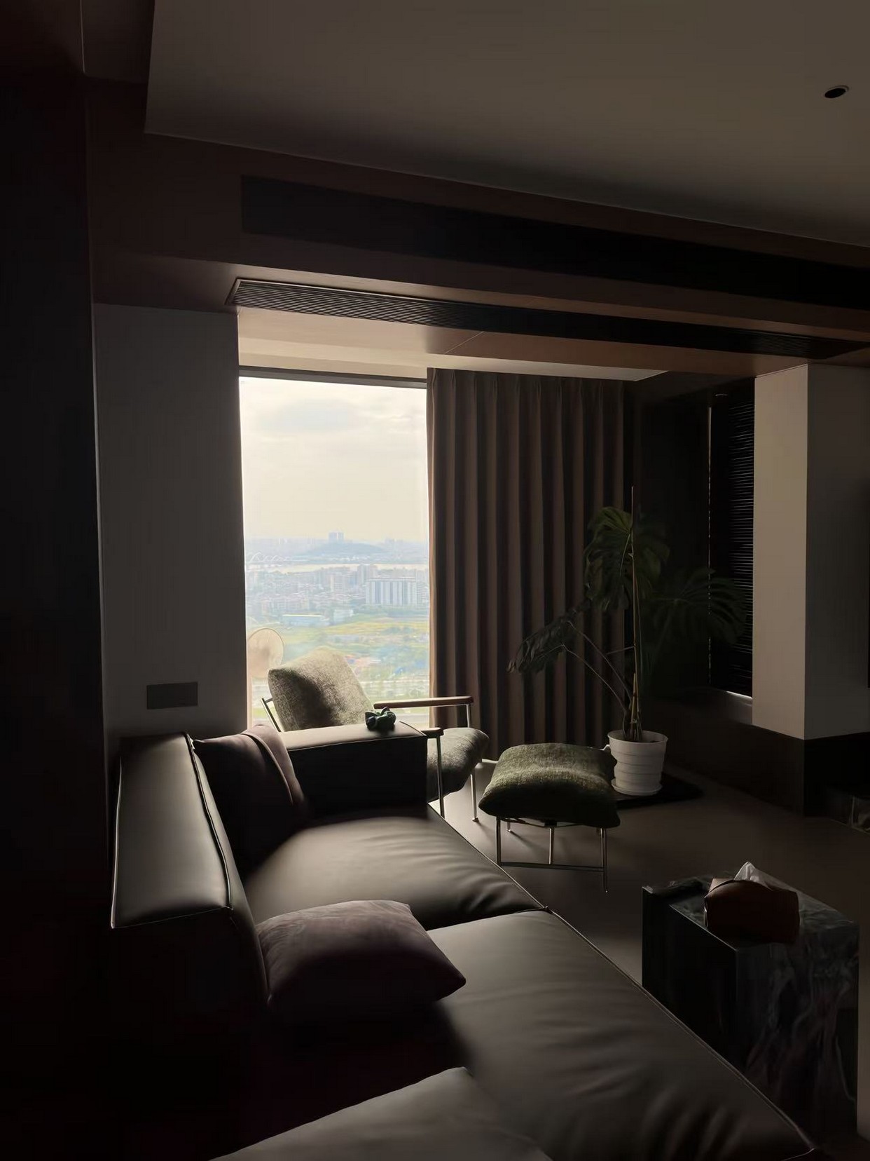A modern living room with a large window, dark curtains, a leather sofa, and a view of a city skyline.